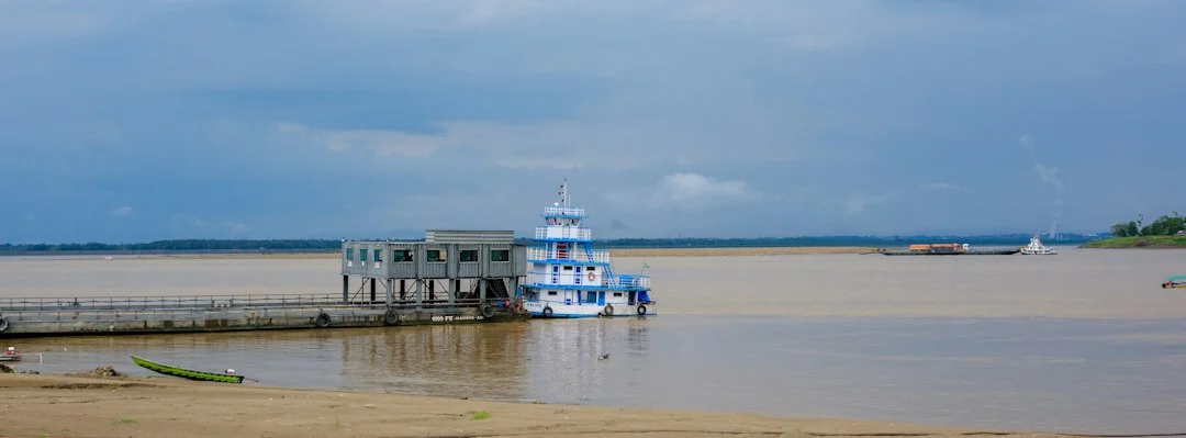 Gebäude auf einem Steg an einem breiten, schlammig braunen Fluss, davor ein festgemachtes Boot