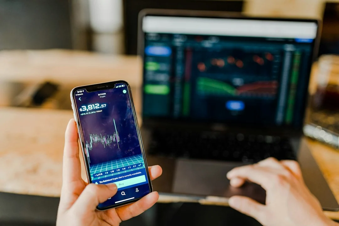 Person hält ein Smartphone und arbeitet an einem Laptop mit Finanzdaten