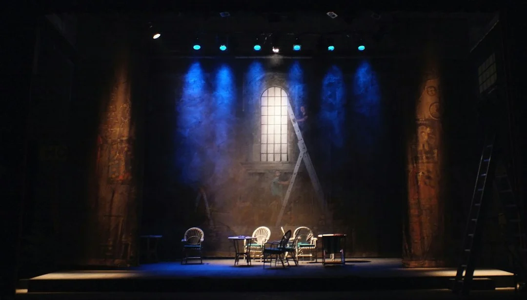 Theaterbühne mit Stühlen und Tischen unter blauem Scheinwerferlicht vor einem bemalten Hintergrund